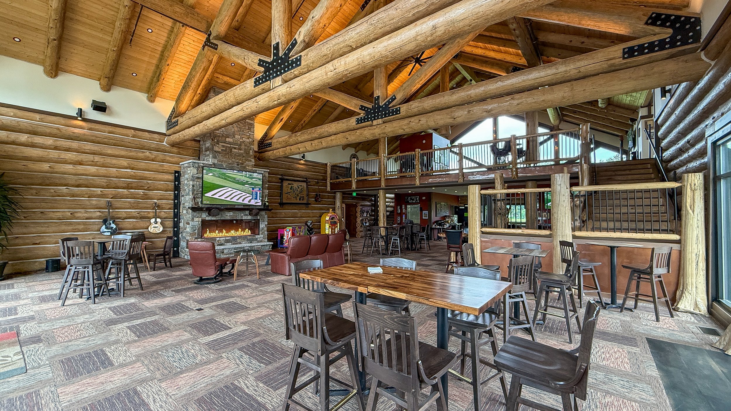 The great room of the log cabin clubhouse, with a stone fireplace, table seating and a balcony above