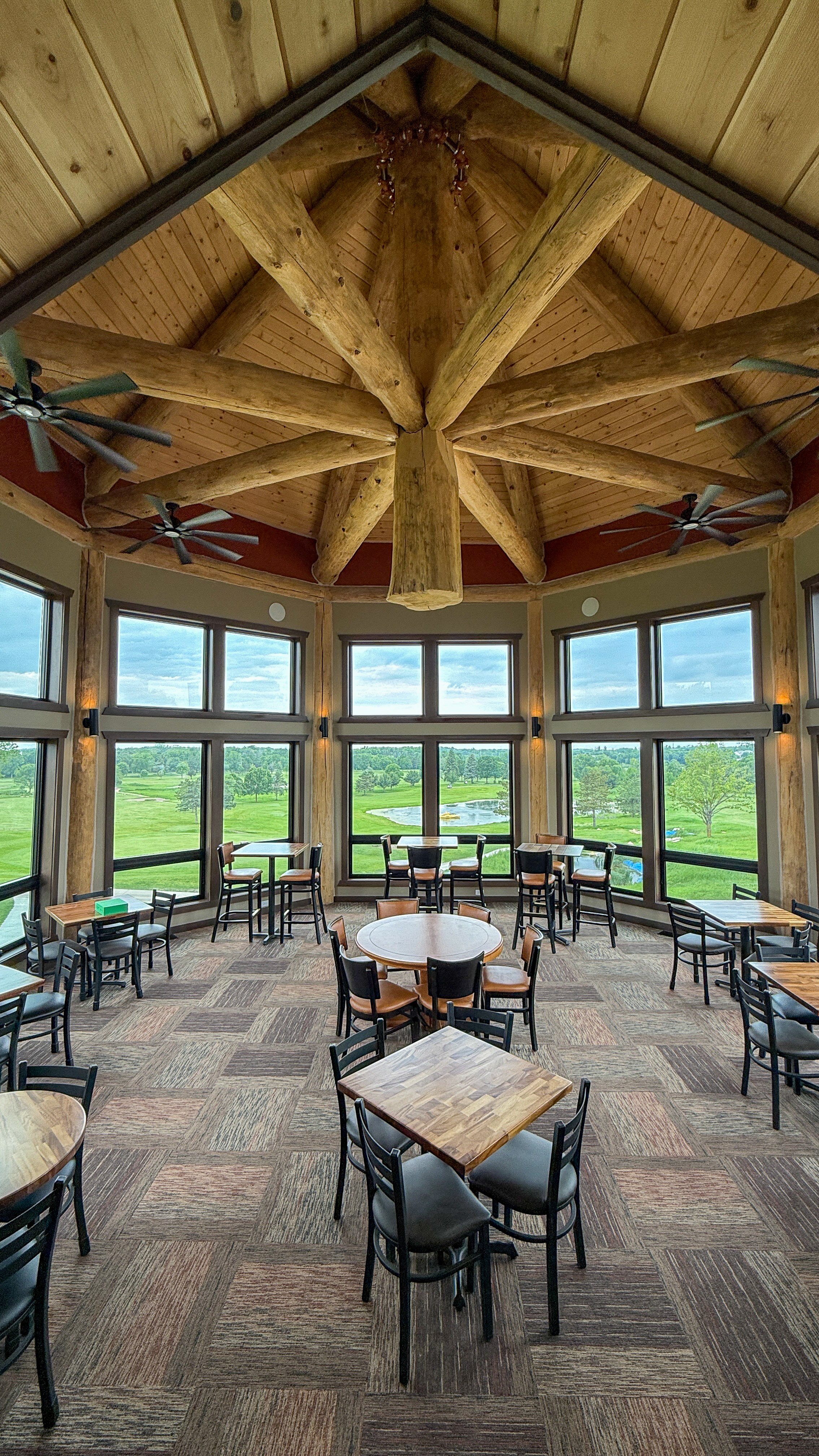 Tables in a windowed room under a vaulted log ceiling, looking out over the course